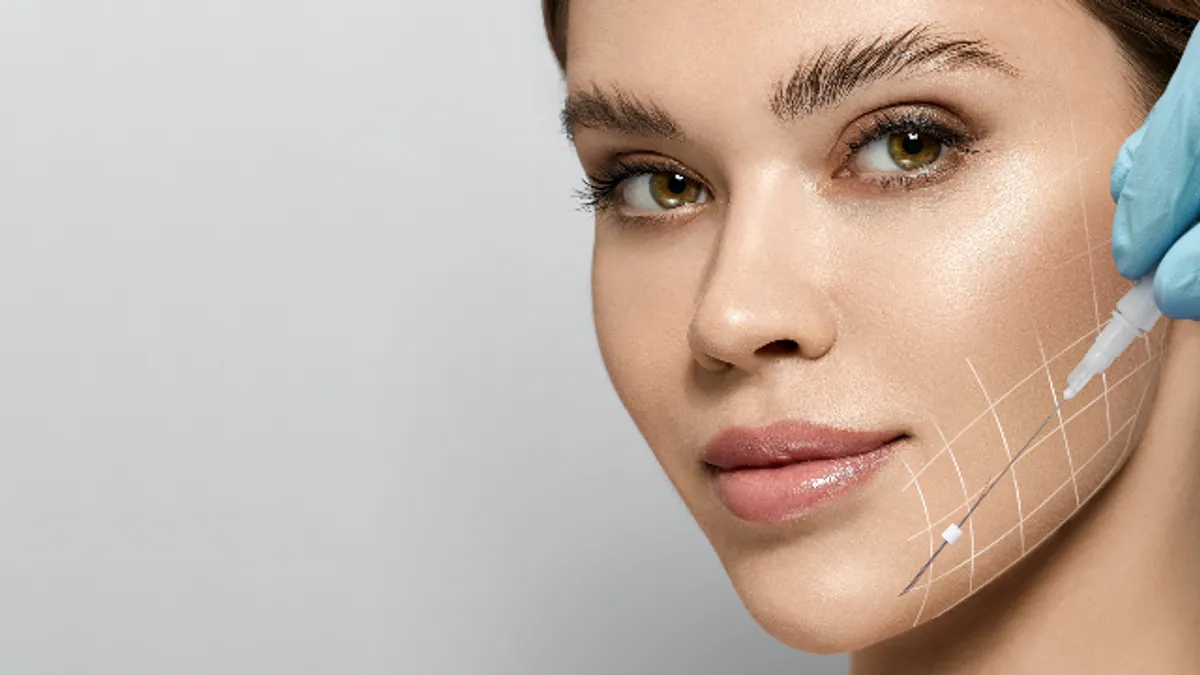 A close-up of a young woman's face with a white grid pattern overlaid on her cheek, while a gloved hand uses a medical tool to demonstrate a facial mapping or treatment area.