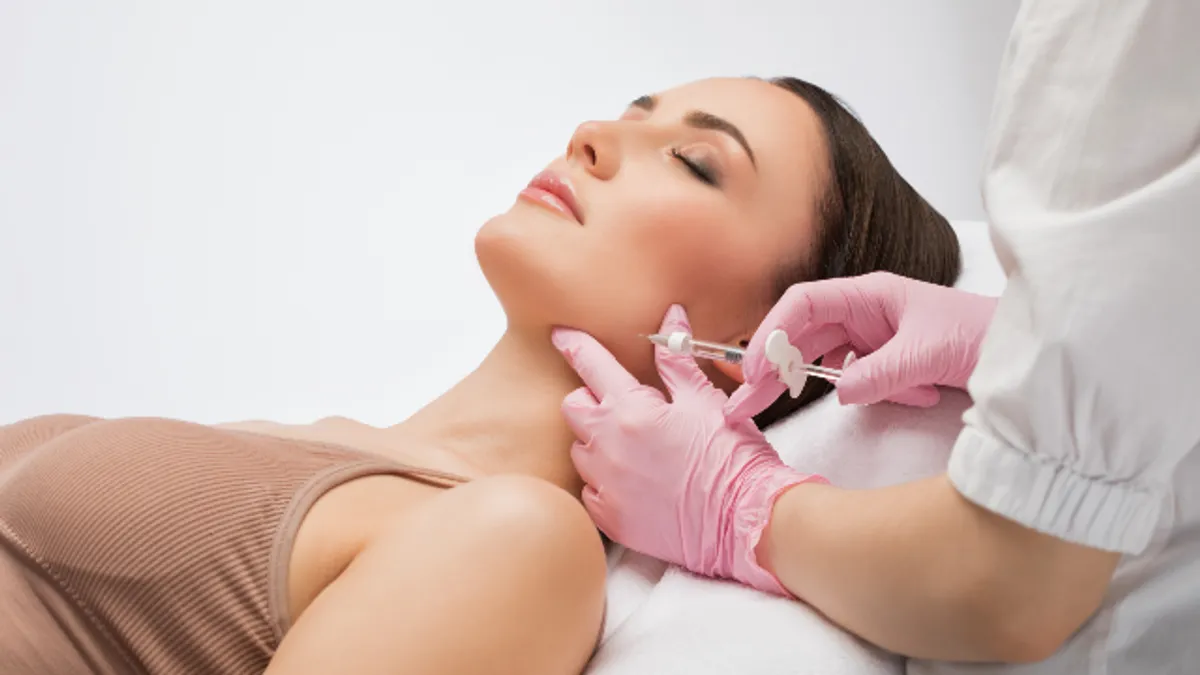 A professional in pink gloves administering a cosmetic injection into the jawline of a young woman lying down with her eyes closed.