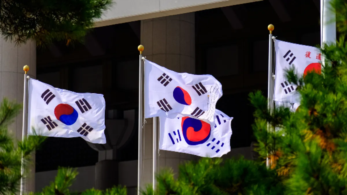 석조 기둥이 있는 건물 앞, 푸른 소나무 가지 사이로 여러 개의 태극기가 깃대에서 휘날리고 있습니다.