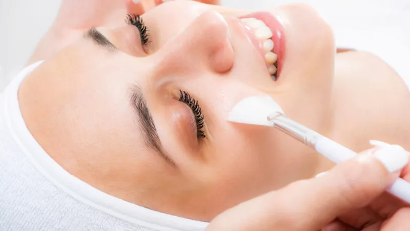 Practitioner applying a facial treatment with a brush near a woman’s cheek.