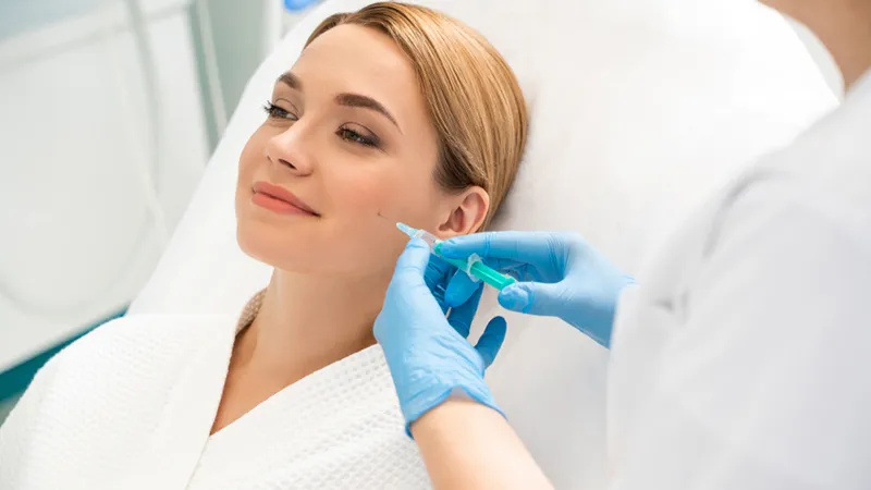 Practitioner holding a syringe near a reclining woman’s cheek.
