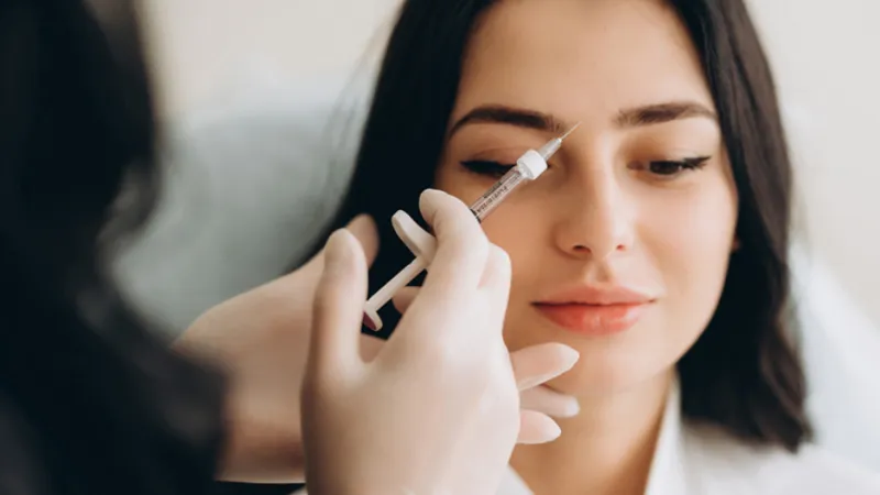Practitioner holding a syringe near a woman’s eyebrow.