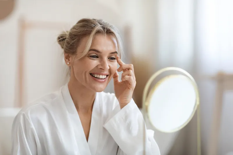 Woman touching the outer corner of her eye while looking in a mirror.