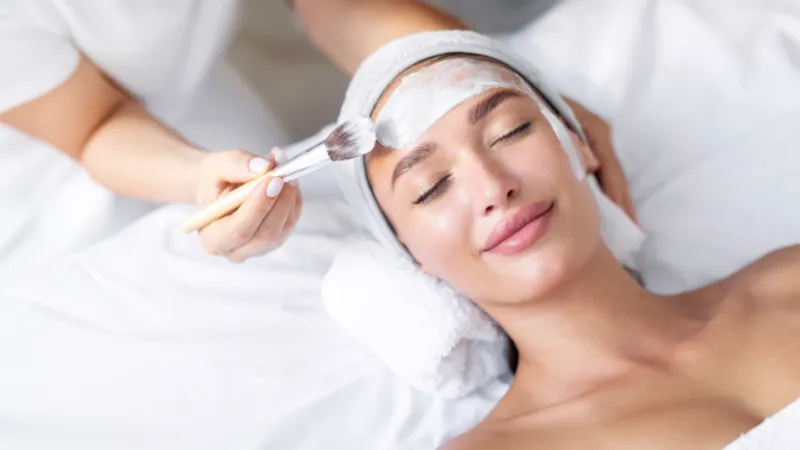 Practitioner applying a facial mask with a brush to a reclining woman.