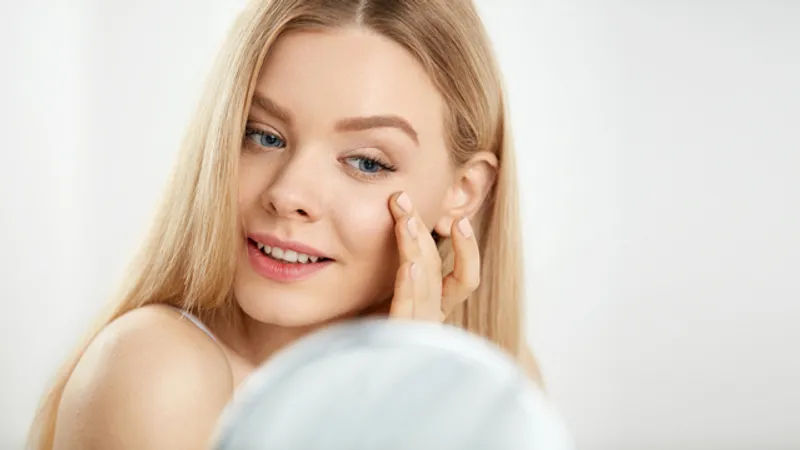 Woman looking in a mirror and examining the appearance of dark circles under her eyes.
