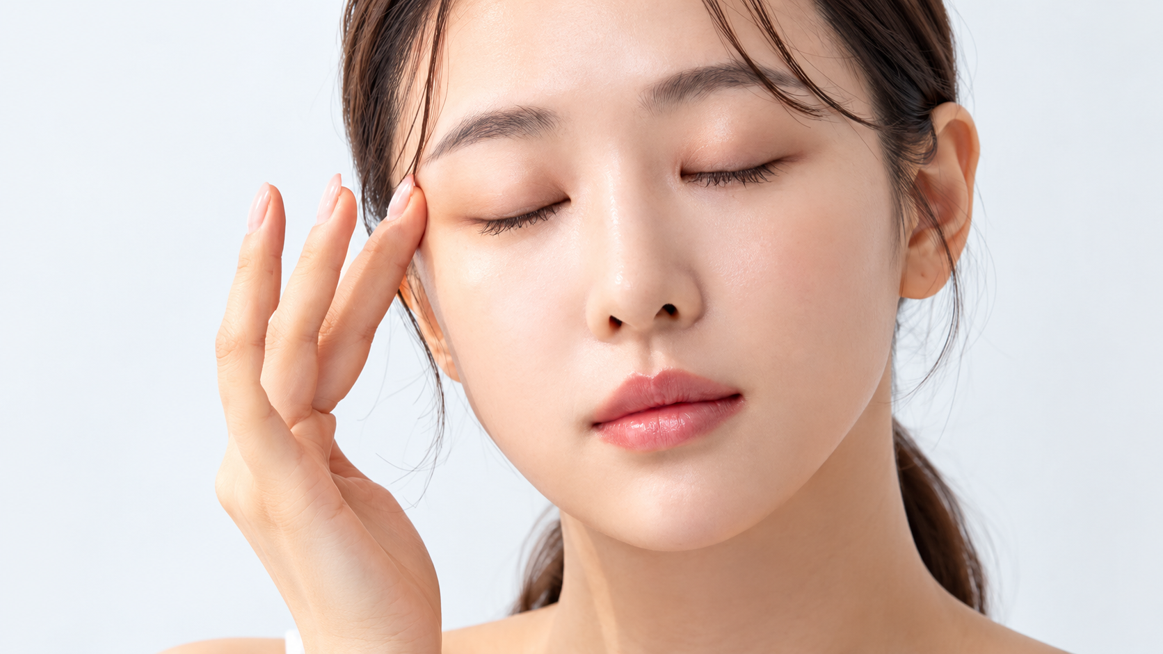 Woman with closed eyes gently touching her temple against a light background.