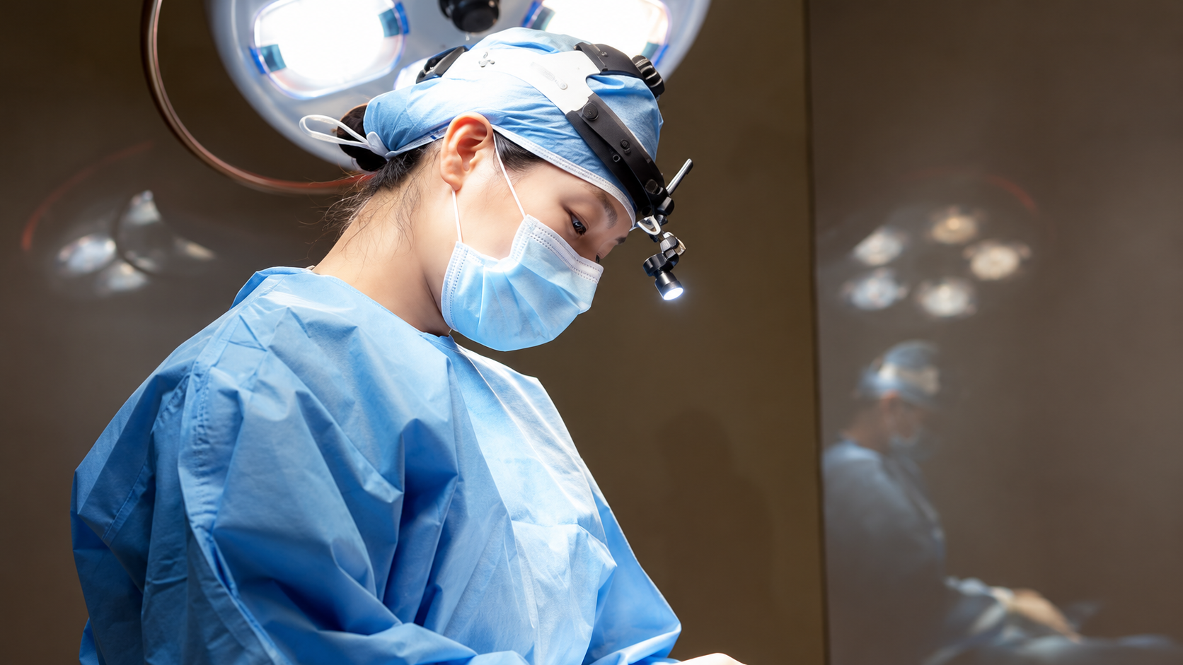 Medical professional in surgical scrubs wearing a headlamp under operating room lights.