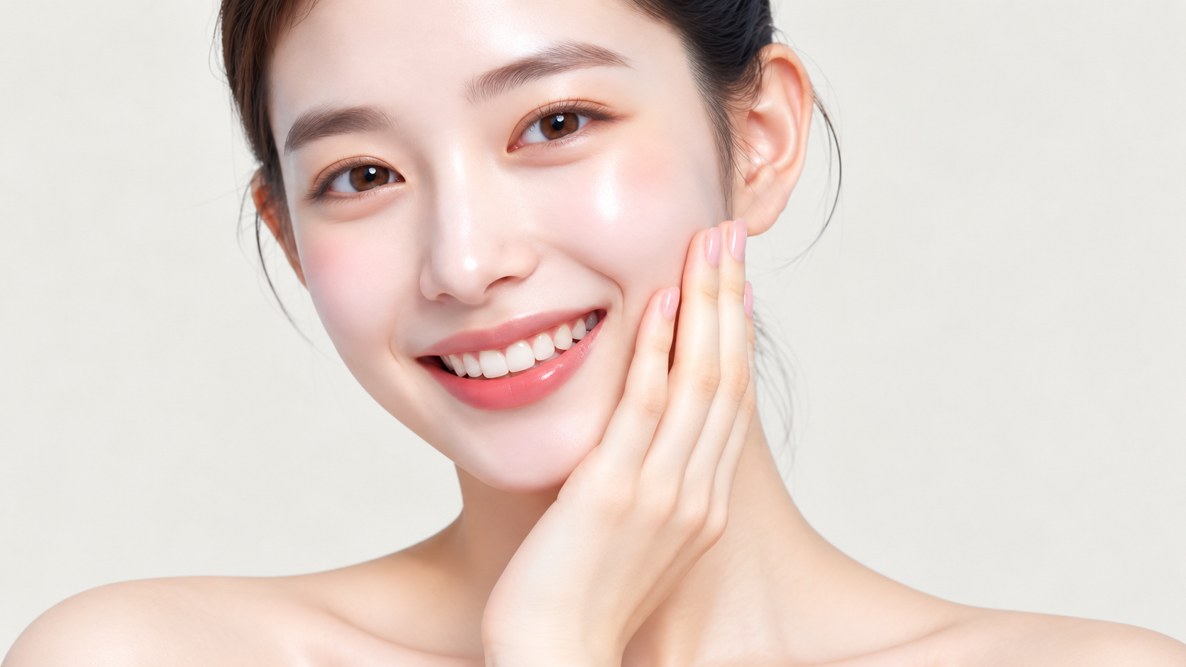 Smiling woman gently touching her cheek while facing the camera against a light background.