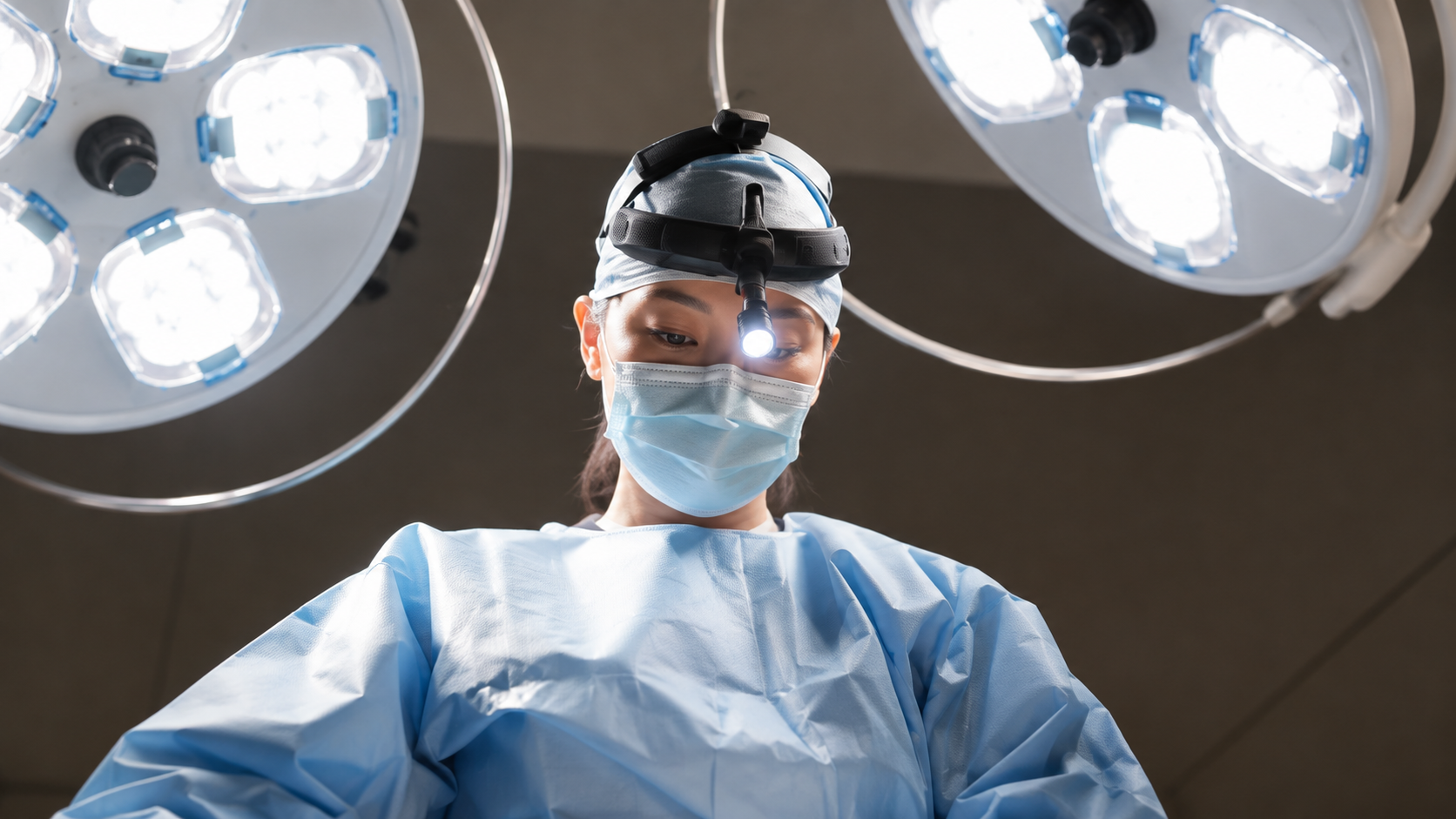 Surgeon wearing a mask and headlight beneath operating room lights.