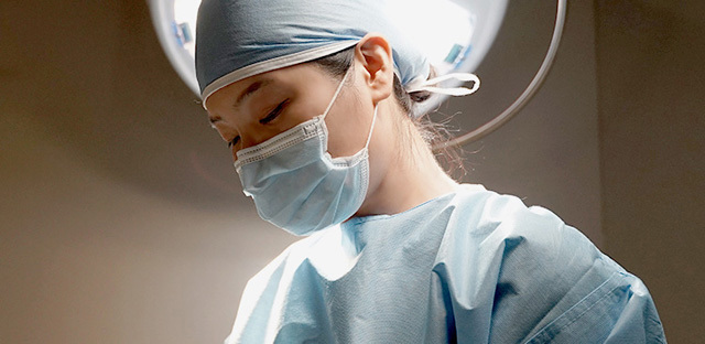 Surgeon wearing a mask and gown beneath operating room lights.