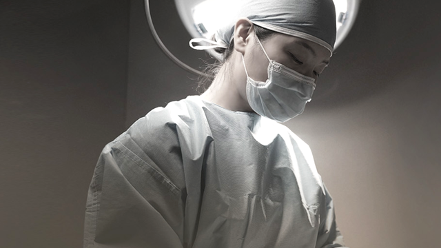 Medical professional wearing a surgical mask and gown under operating lights.